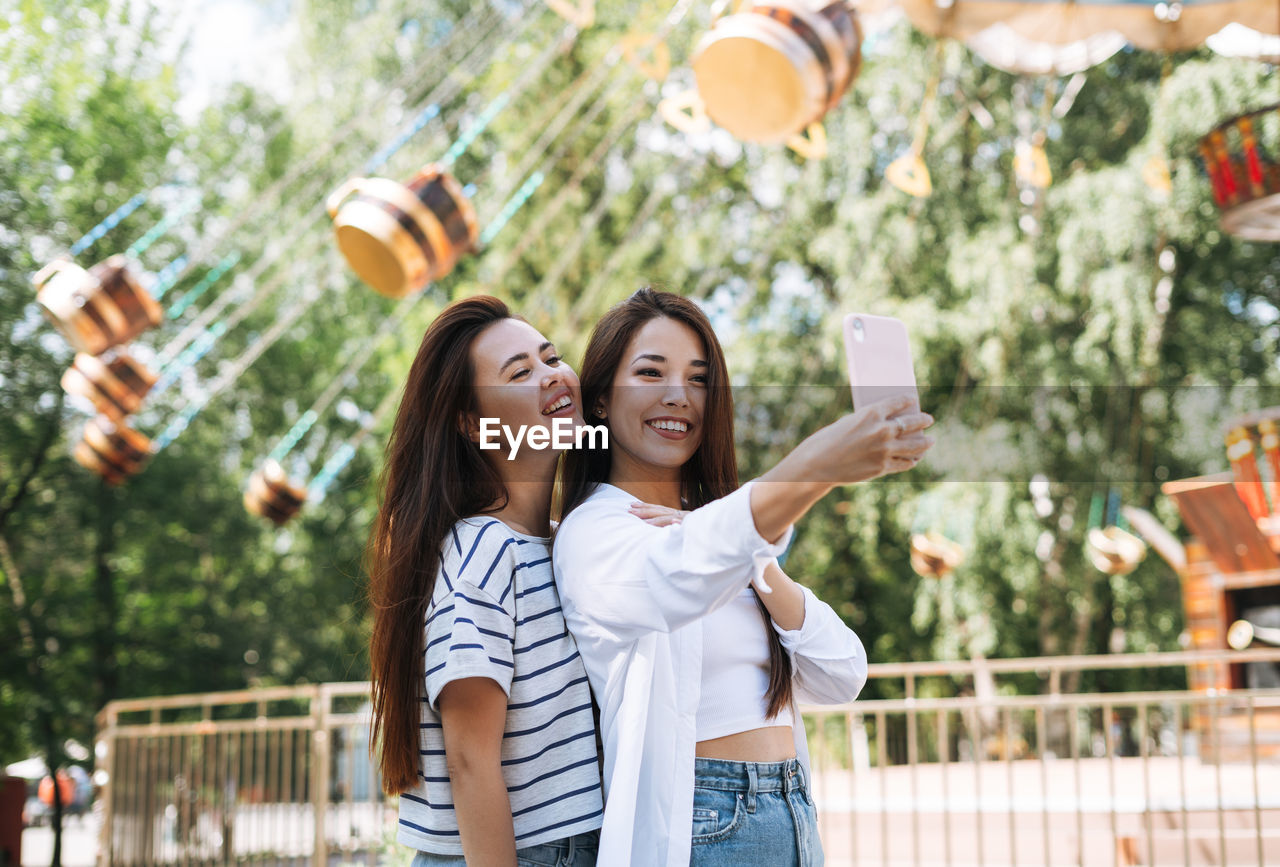 Young women with long hair friends having fun taking selfie on mobile phone at amusement park