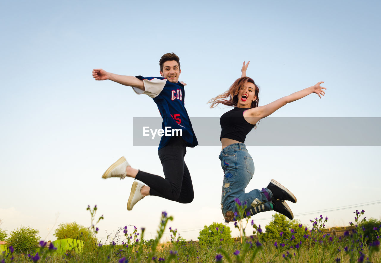 Full length side view of excited young couple jumping against sky on field