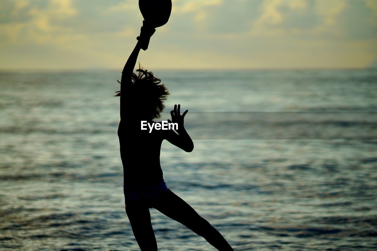 Silhouette boy with hand raised jumping by sea against sky during sunset