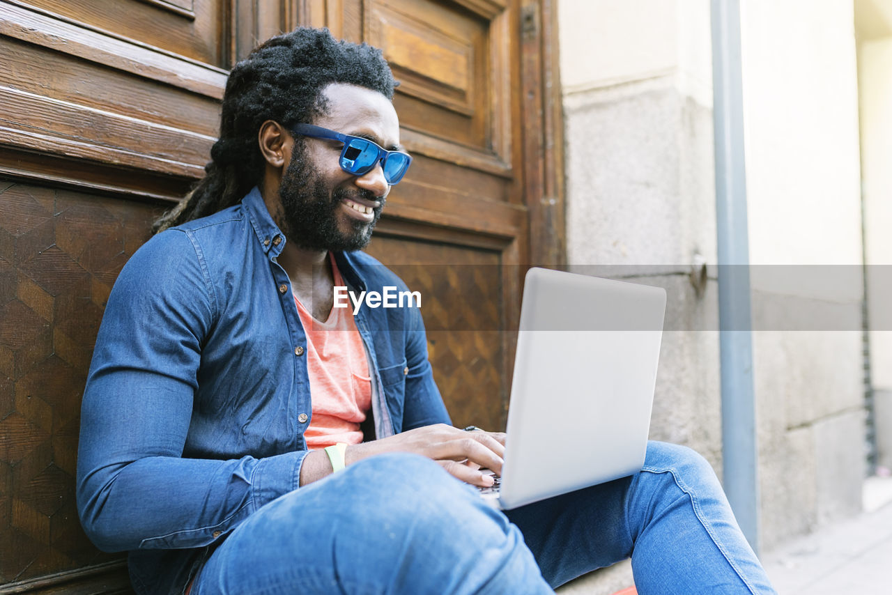 Smiling young man using laptop while sitting in city