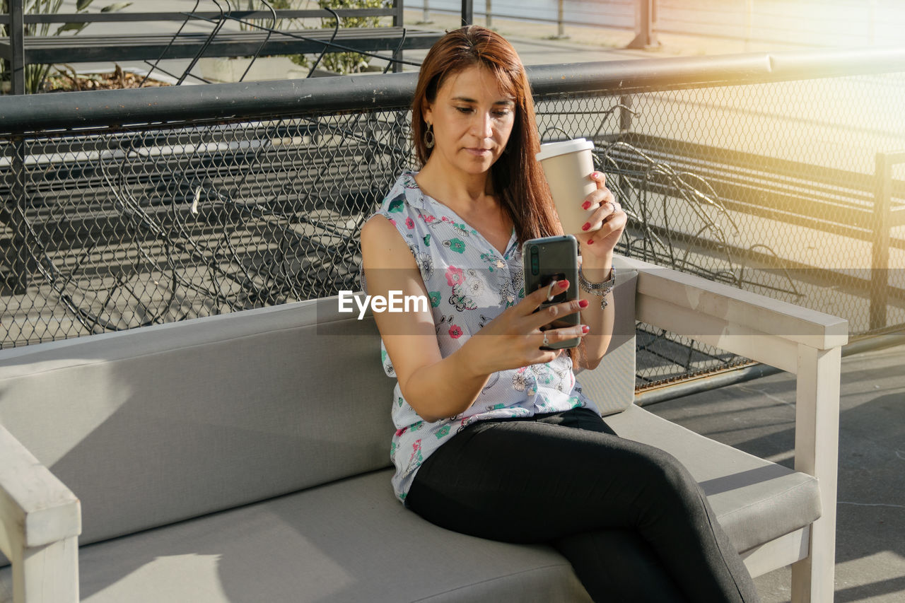 Hispanic woman in coffee shop reading phone sitting on external couch holding cup of coffee