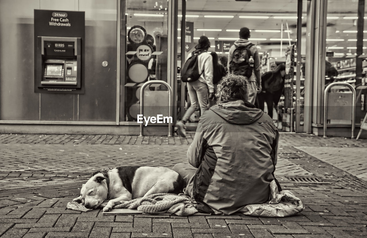 street, road, infrastructure, monochrome, homelessness, dog, men, city, adult, architecture, black and white, black, poverty, footpath, group of people, sitting, person, monochrome photography, relaxation, lifestyles, white, mammal, animal, sidewalk, animal themes, one animal, rail transportation, domestic animals, full length, canine, pet, rear view, women, city life