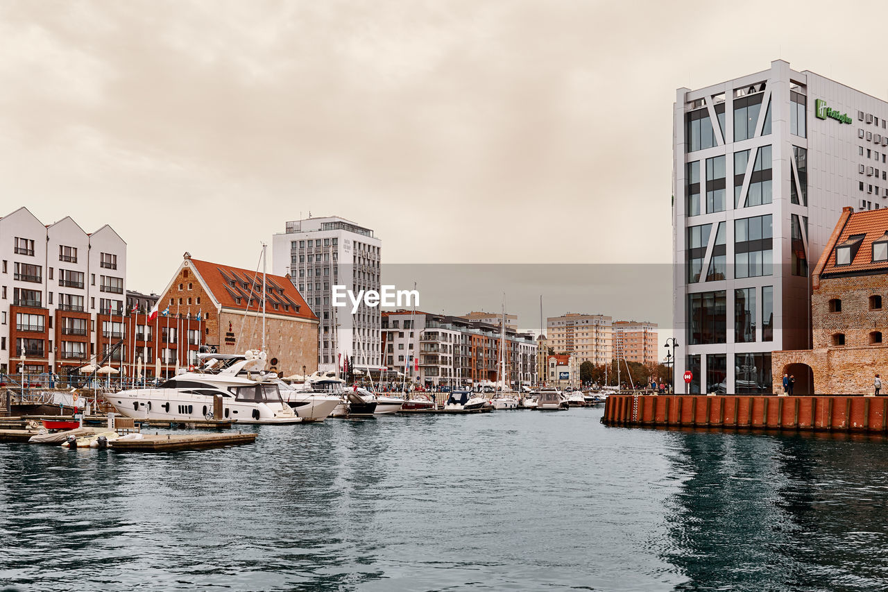 SAILBOATS MOORED IN RIVER BY BUILDINGS AGAINST SKY