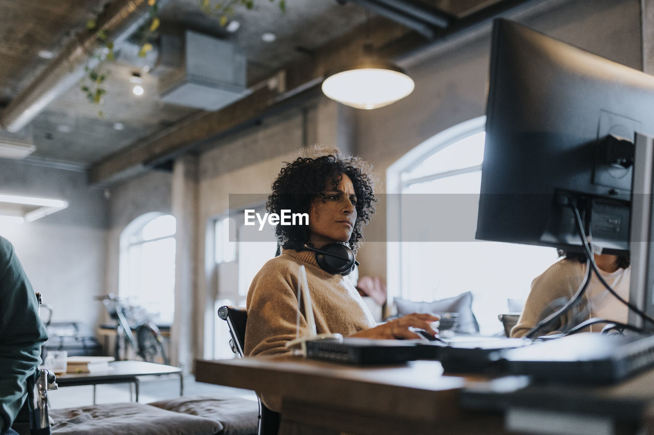 Female hacker with wireless headphones around neck using computer while working in office