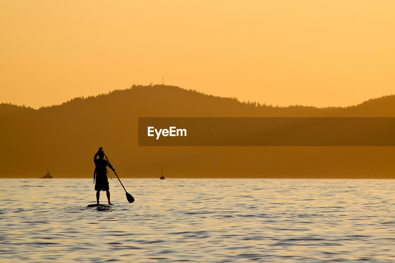Silhouette person on paddleboard in river against mountains during sunset