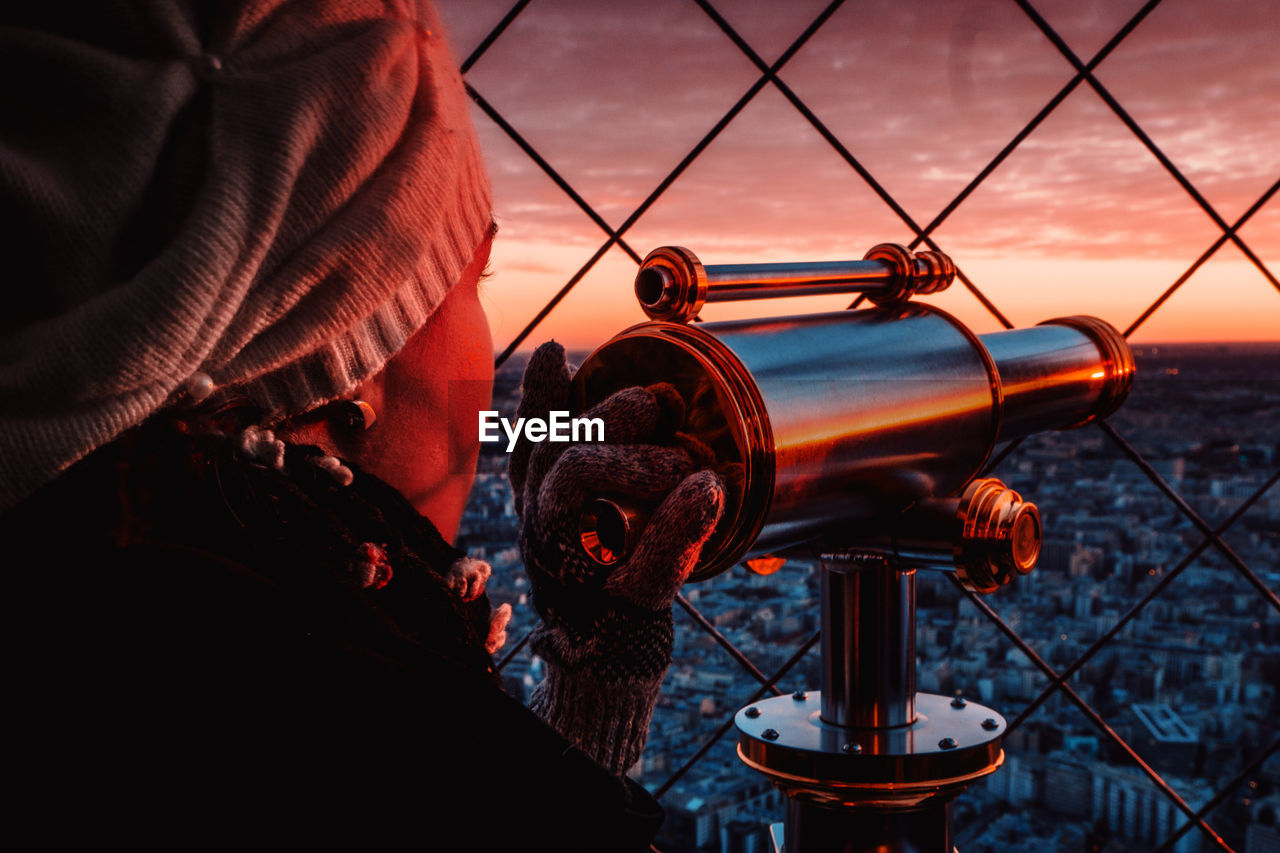 Woman holding coin operated binoculars by tour eiffel against sky during sunset