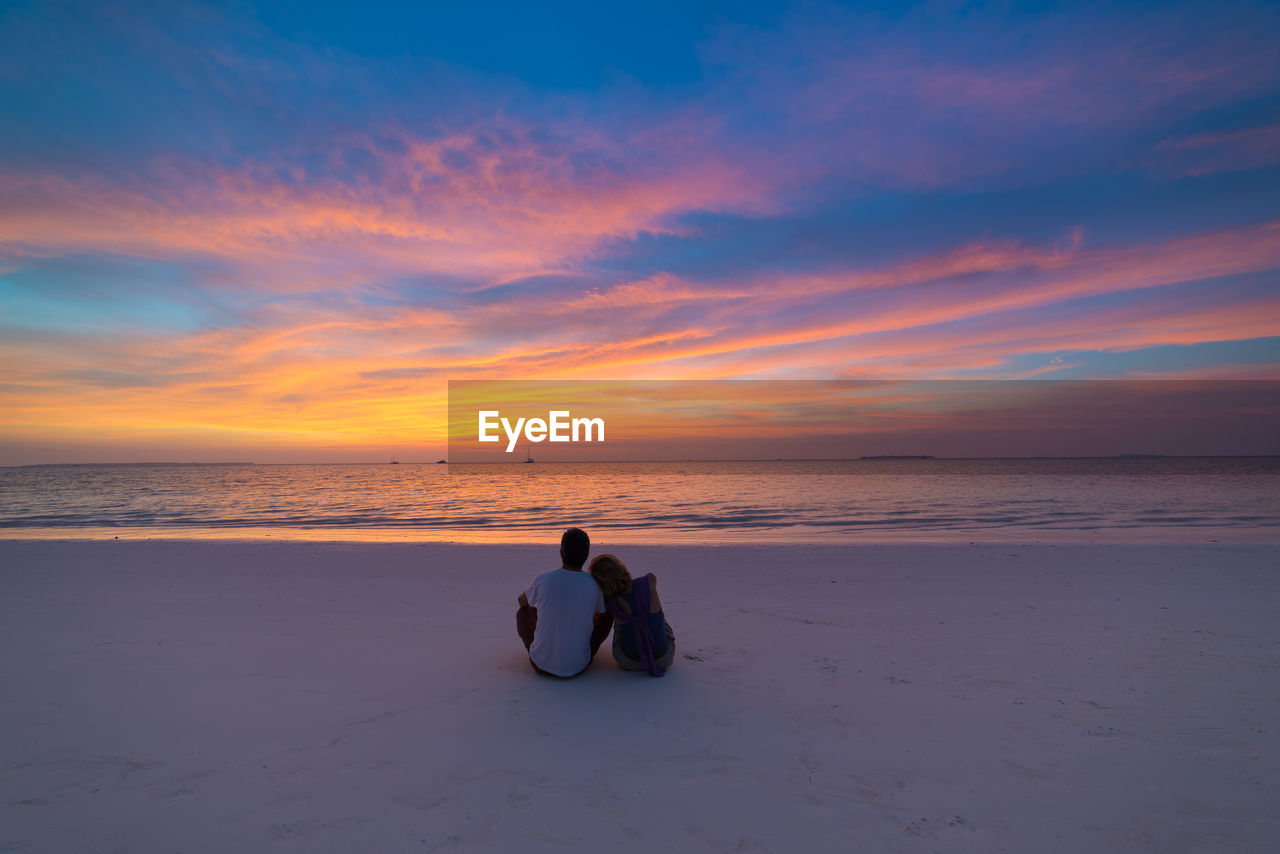 Rear view of couple sitting at beach against sky during sunset