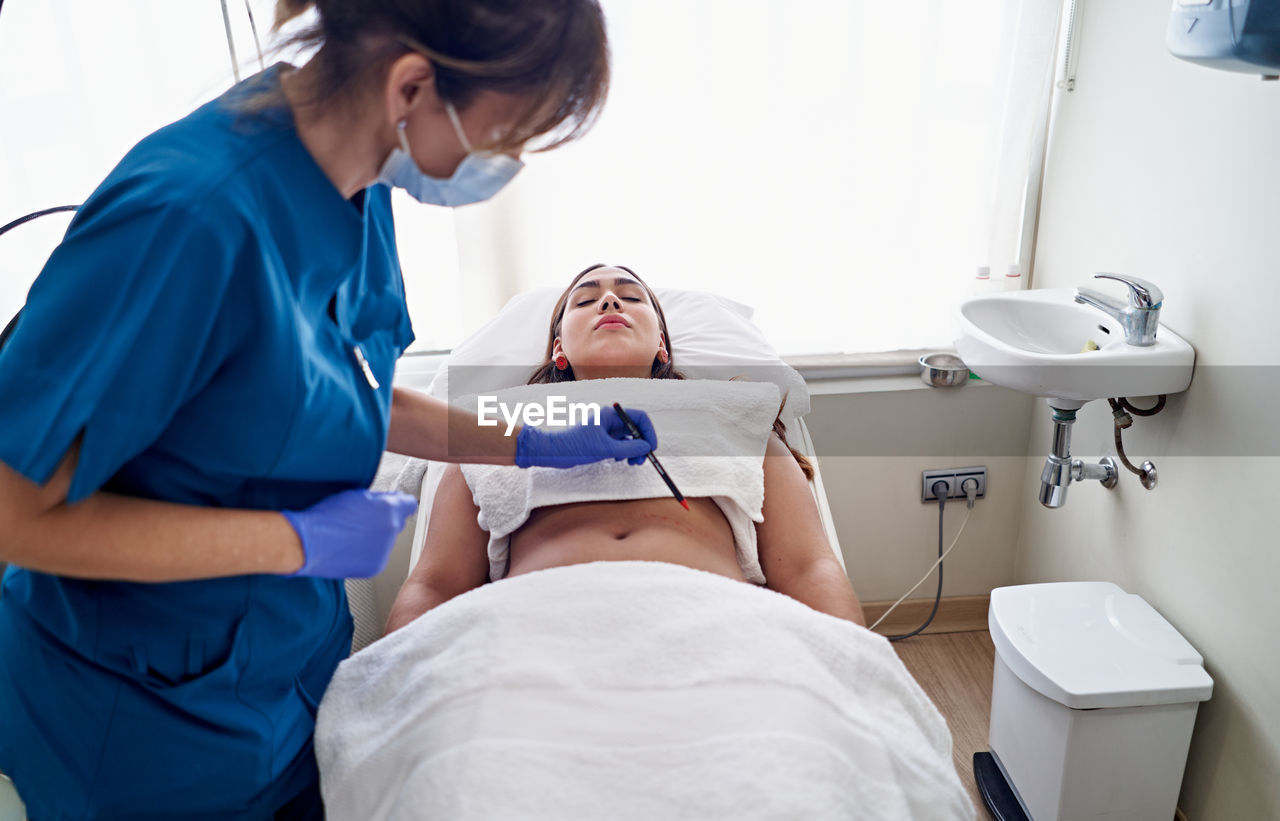 From above beautician in uniform and face mask drawing with pencil preparing to do laser lipolysis to female client during work in aesthetic clinic