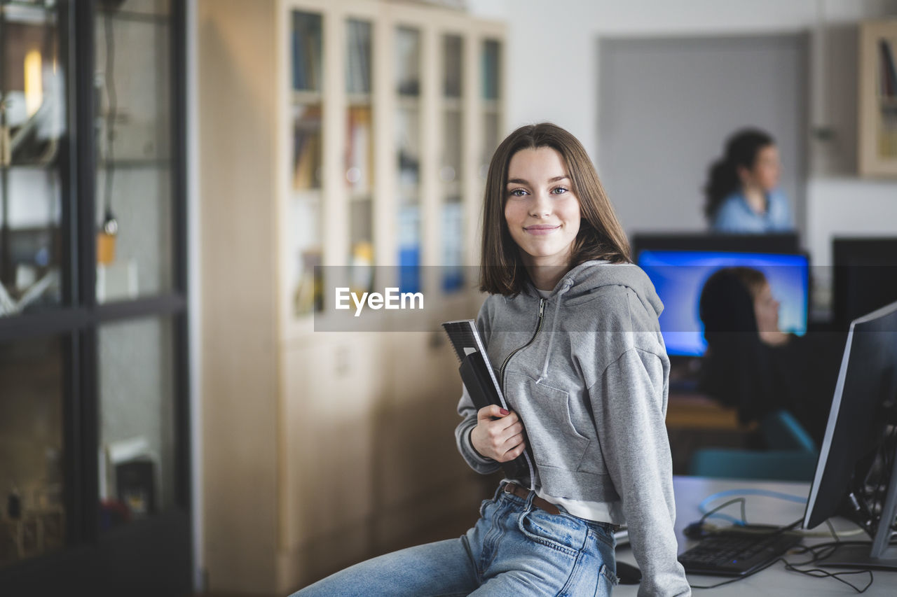 Portrait of smiling high school female student sitting with books on desk in classroom