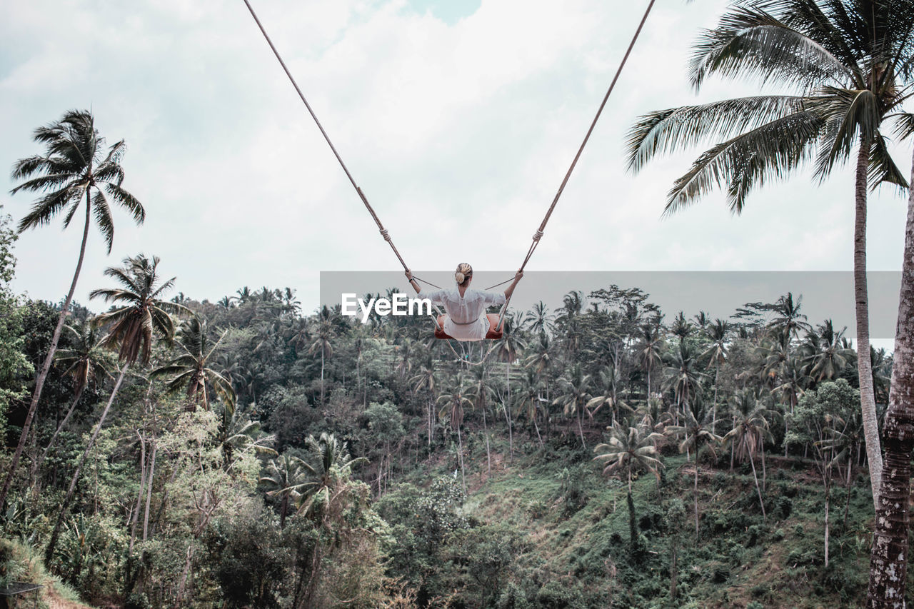 Woman sitting on swing by palm trees against sky