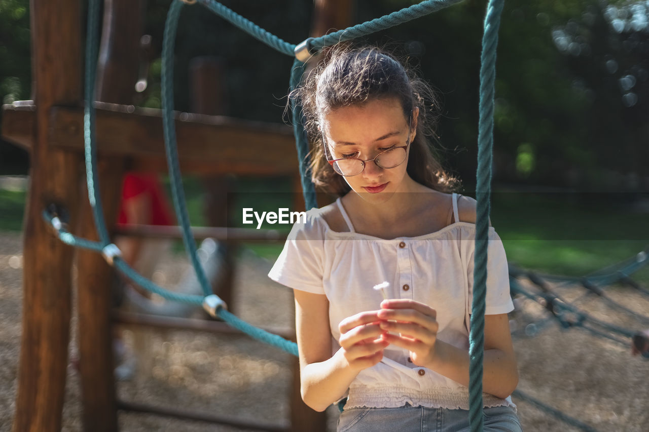 A caucasian teenage girl sits on a rope swing and looks thoughtfully and at a daisy in her hands