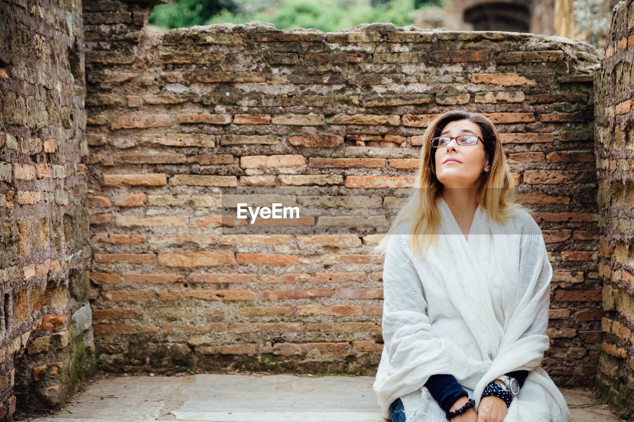 Young woman looking away while sitting against brick wall