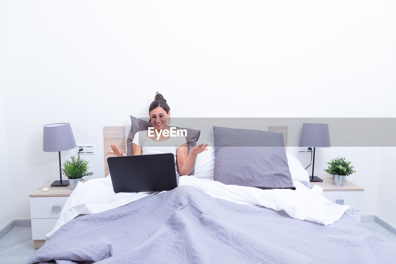 Happy smiling young woman relaxing in her bed with laptop computer. opening arms surprised