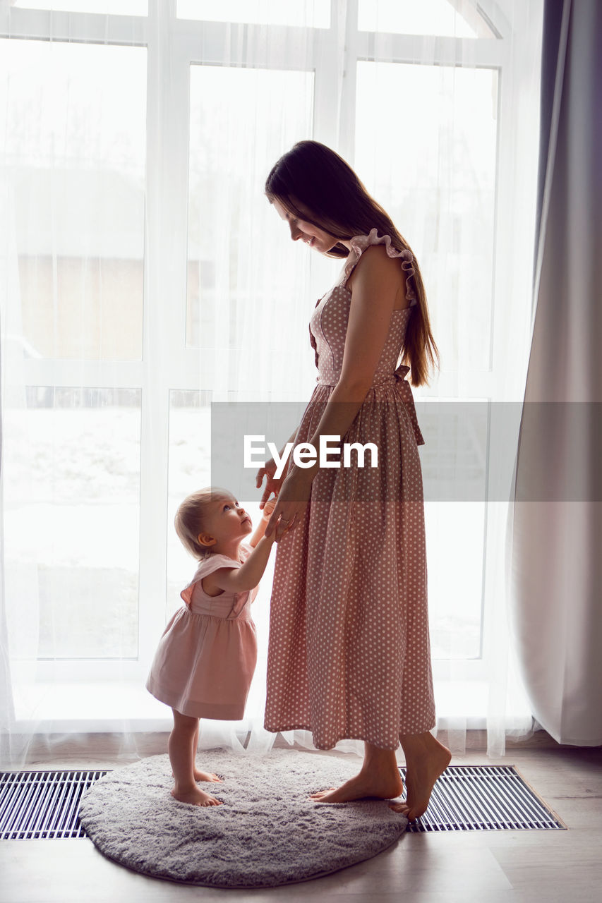 A young woman stands at a large window with a girl child in pink dresses at home barefoot