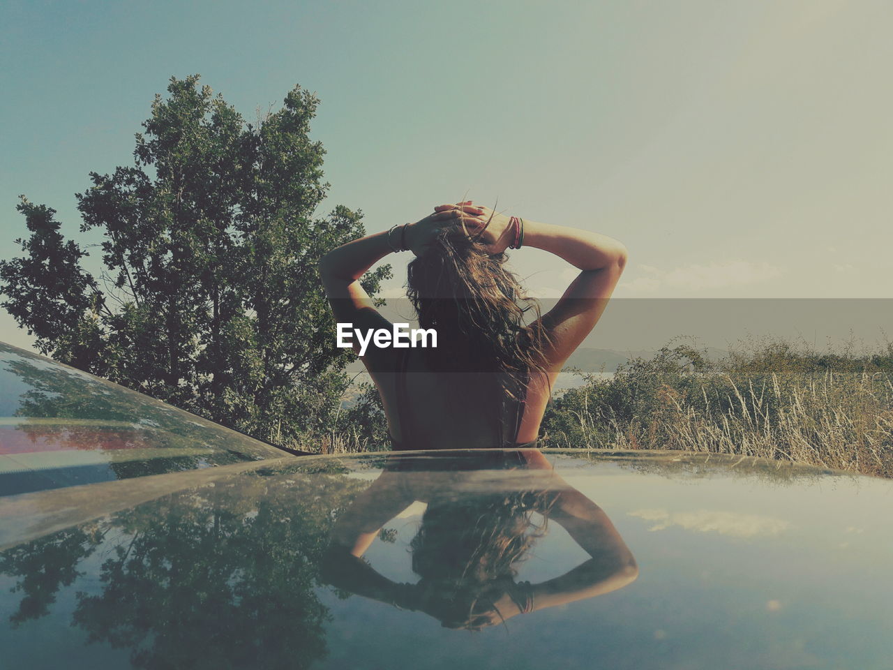 Rear view of woman with hands behind head standing by car against sky