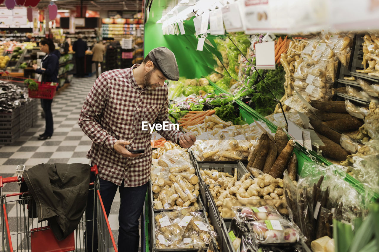 Man holding smart phone while buying root vegetable in supermarket