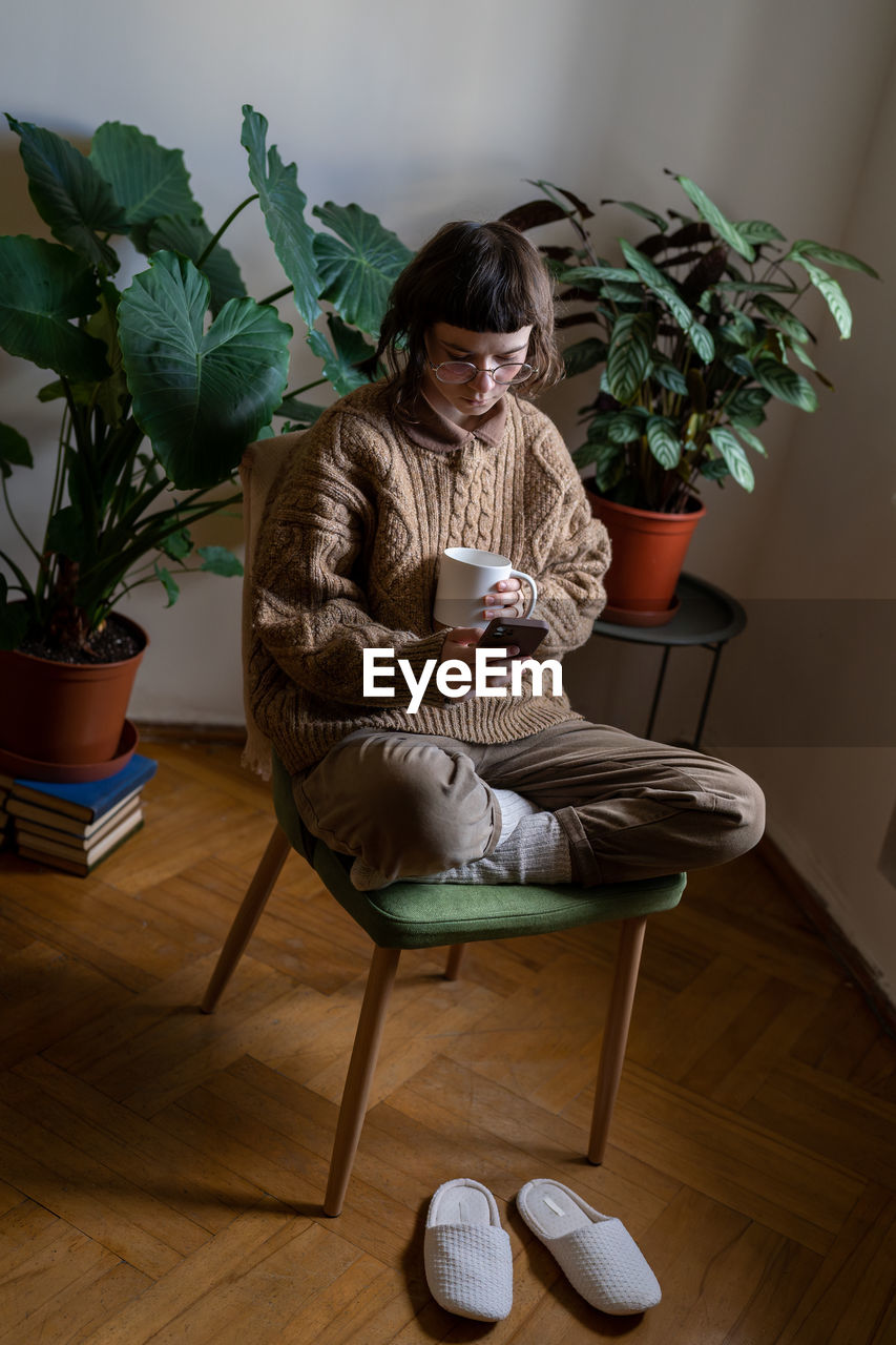 Teen girl sitting on chair with cup of tea, reading news, scrolling photos, messaging in smartphone