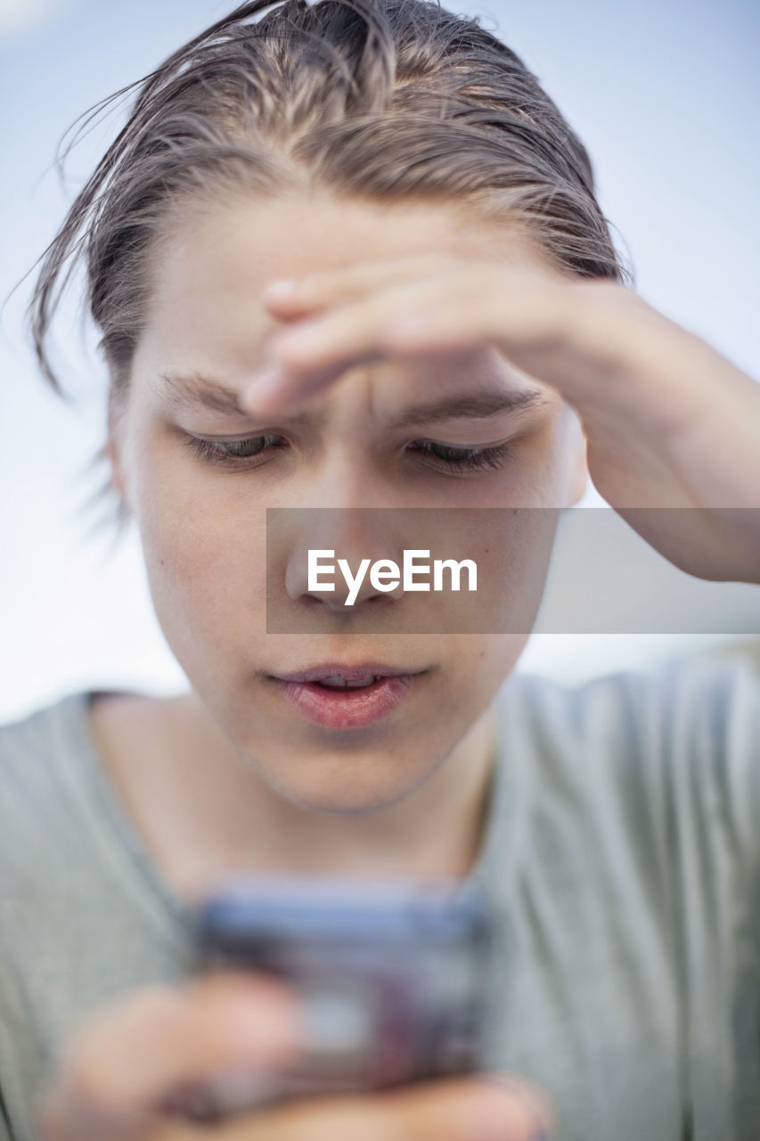 Young man shielding his eyes while using mobile phone against sky