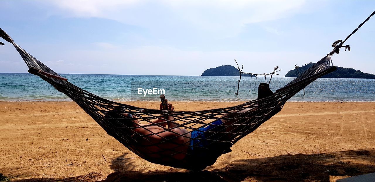 Mature man using mobile phone while relaxing on hammock at beach against sky