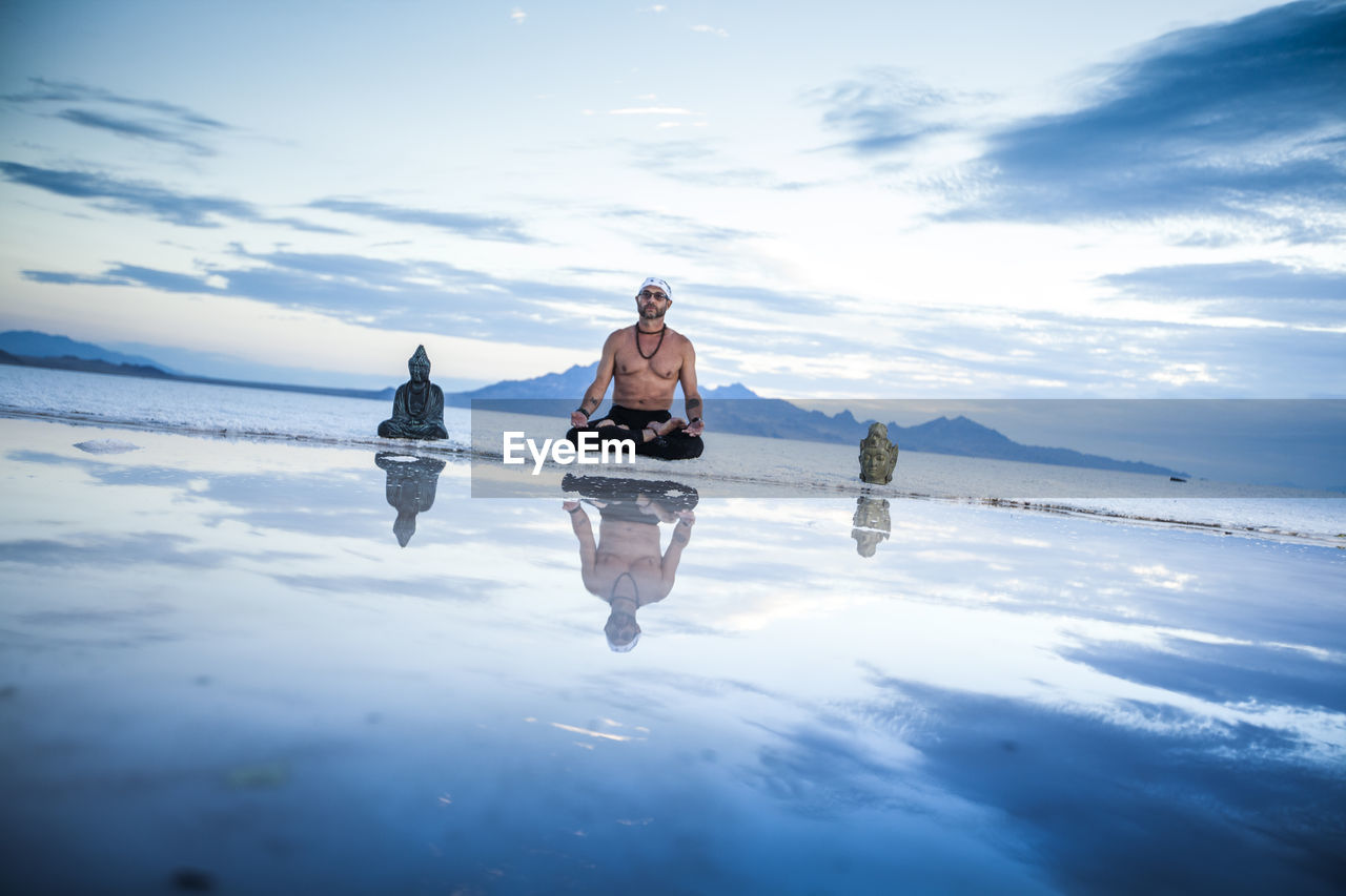 Shirtless mature man meditating while sitting between statues on shore at beach against sky