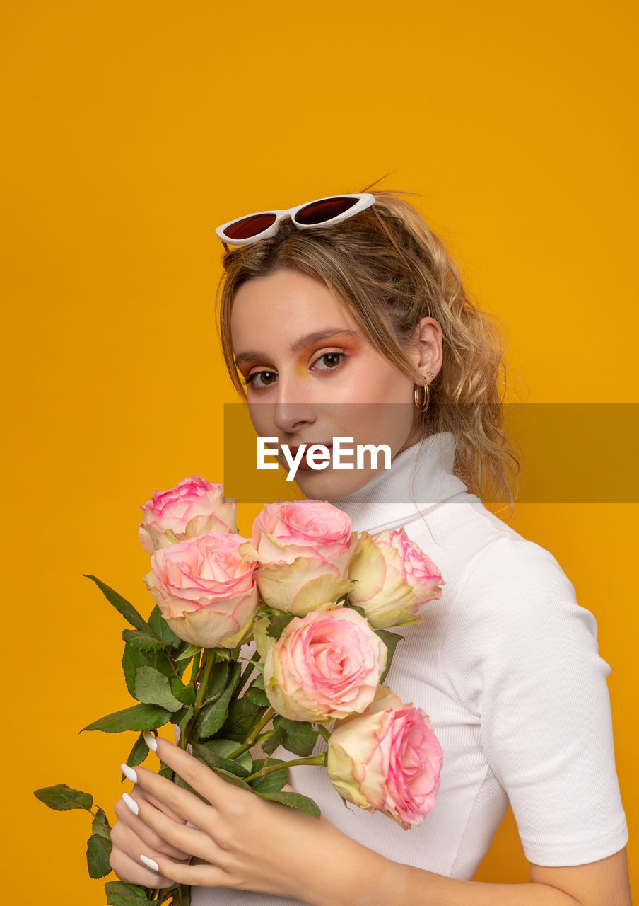 Young beautiful female in white outfit and trendy sunglasses holding delicate pink roses while standing on yellow background in photo studio