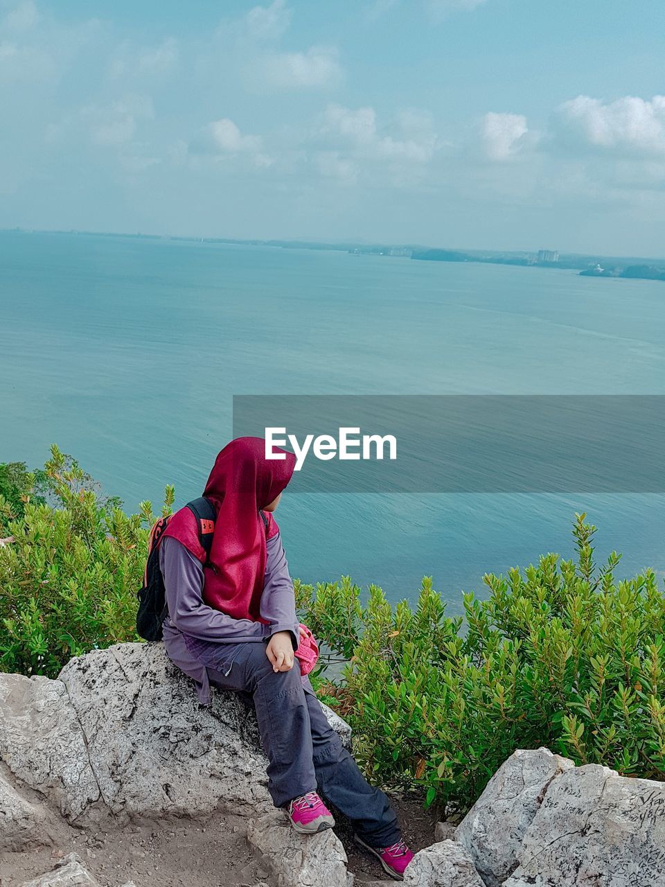 PERSON SITTING ON ROCK BY SEA AGAINST SKY