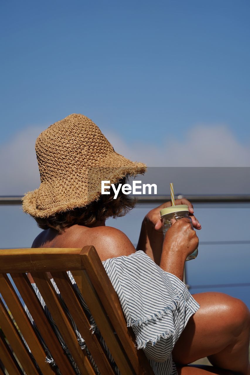 adult, hat, one person, water, sky, relaxation, vacation, trip, nature, leisure activity, clothing, summer, sitting, holiday, sea, women, rear view, sun hat, straw hat, day, lifestyles, holding, food and drink, travel, cowboy hat, seat, men, outdoors, drink, refreshment, chair, sunlight, clear sky, person, travel destinations, land, beach, tourism