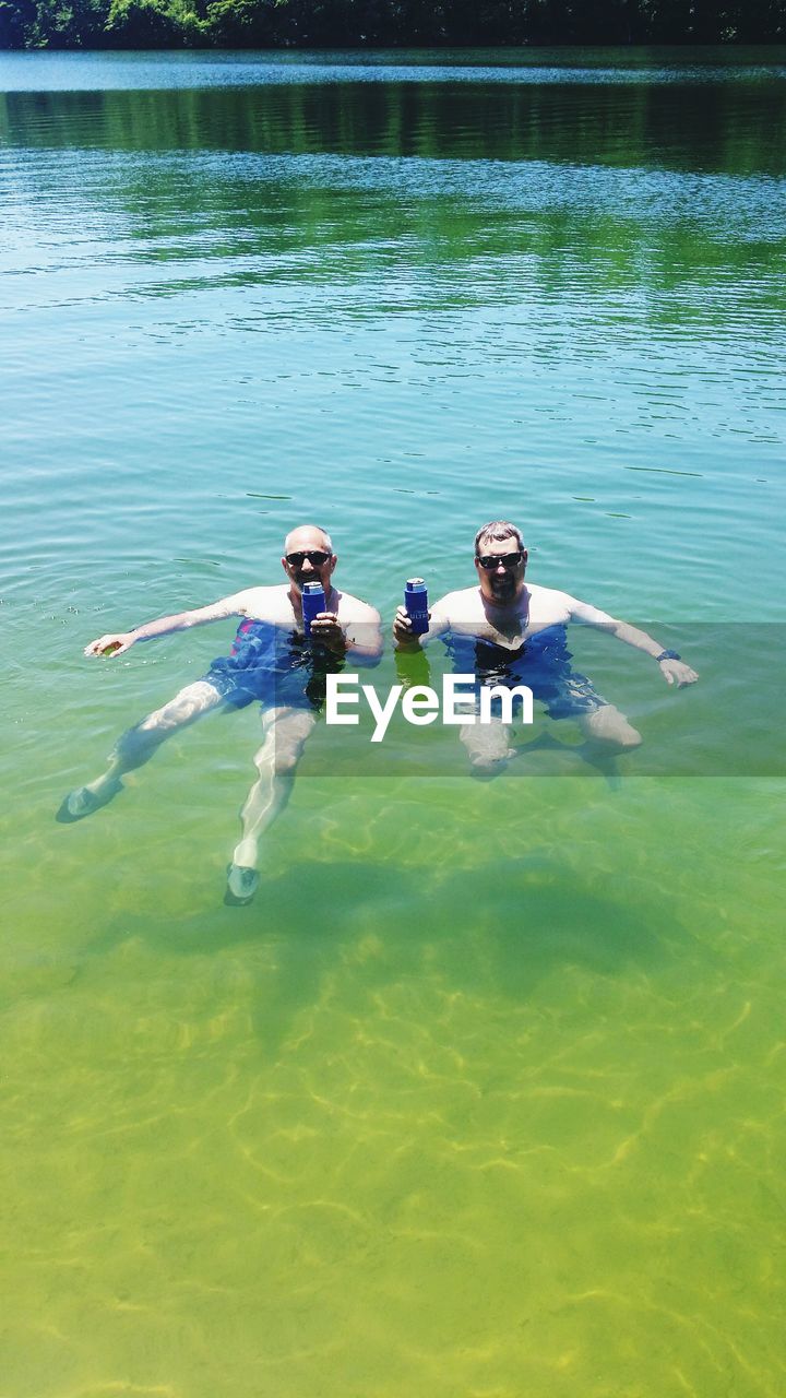 High angle portrait of male friends holding drink cans while swimming in lake