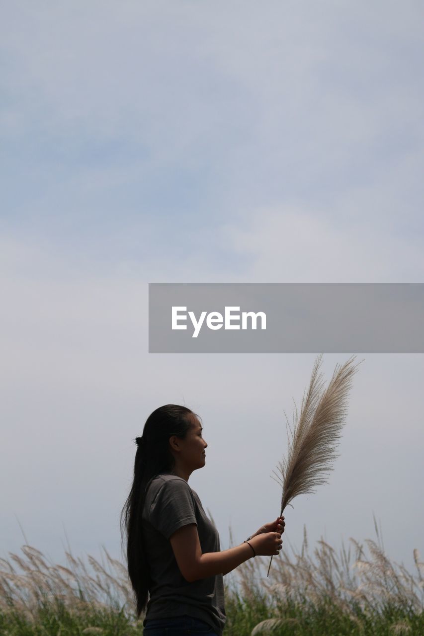 Side view of woman holding pampas grass while standing on field against sky