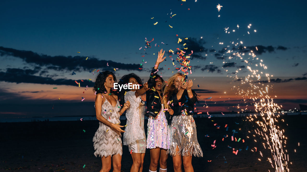 Cheerful young female friends with firework and confetti at beach against sky during sunset