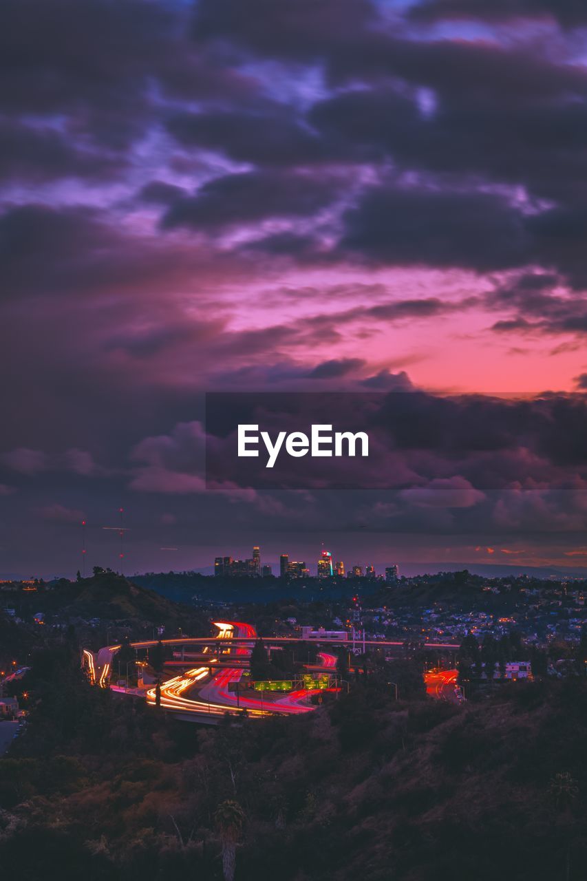 HIGH ANGLE VIEW OF ILLUMINATED CITYSCAPE AGAINST DRAMATIC SKY AT SUNSET