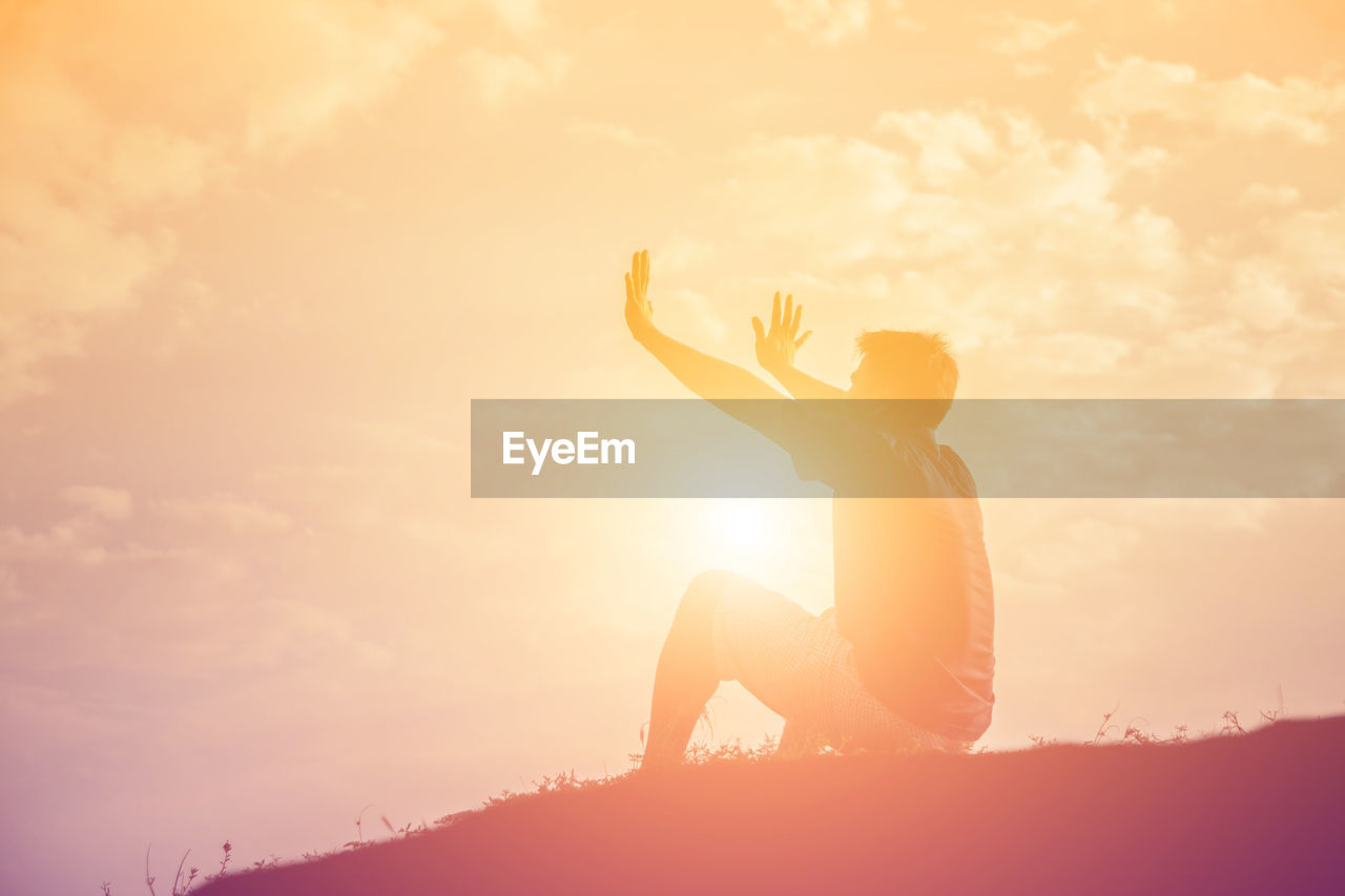 Side view of man gesturing while sitting on land against sky during sunset