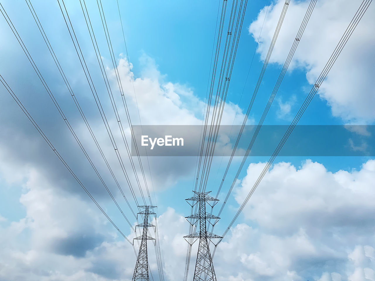 Low angle view of high voltage towers and power lines against blue cloudy sky