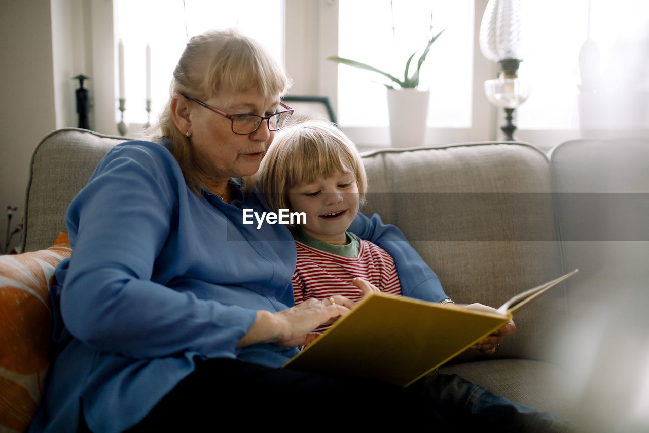 Grandmother reading storybook for grandson while sitting on sofa in living room