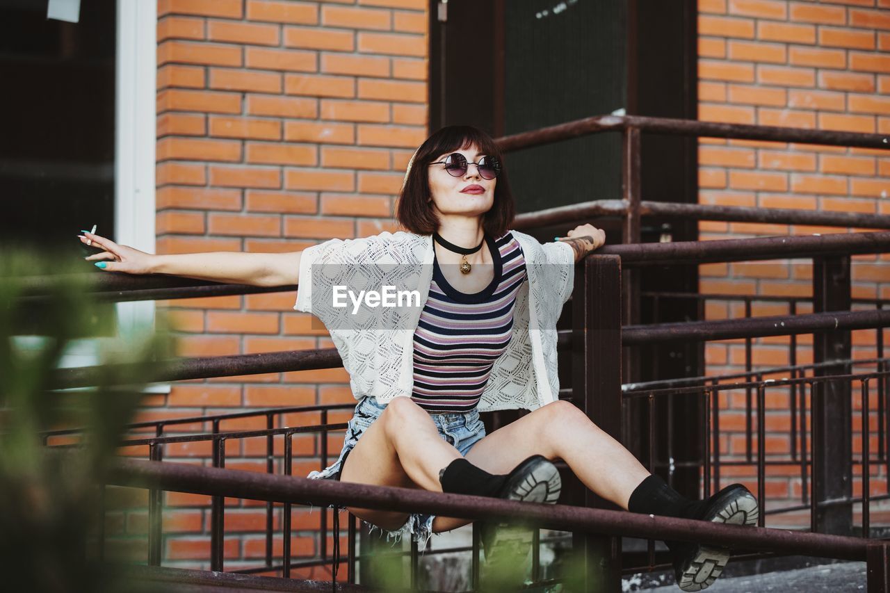 Full length of woman holding cigarette while sitting against railing