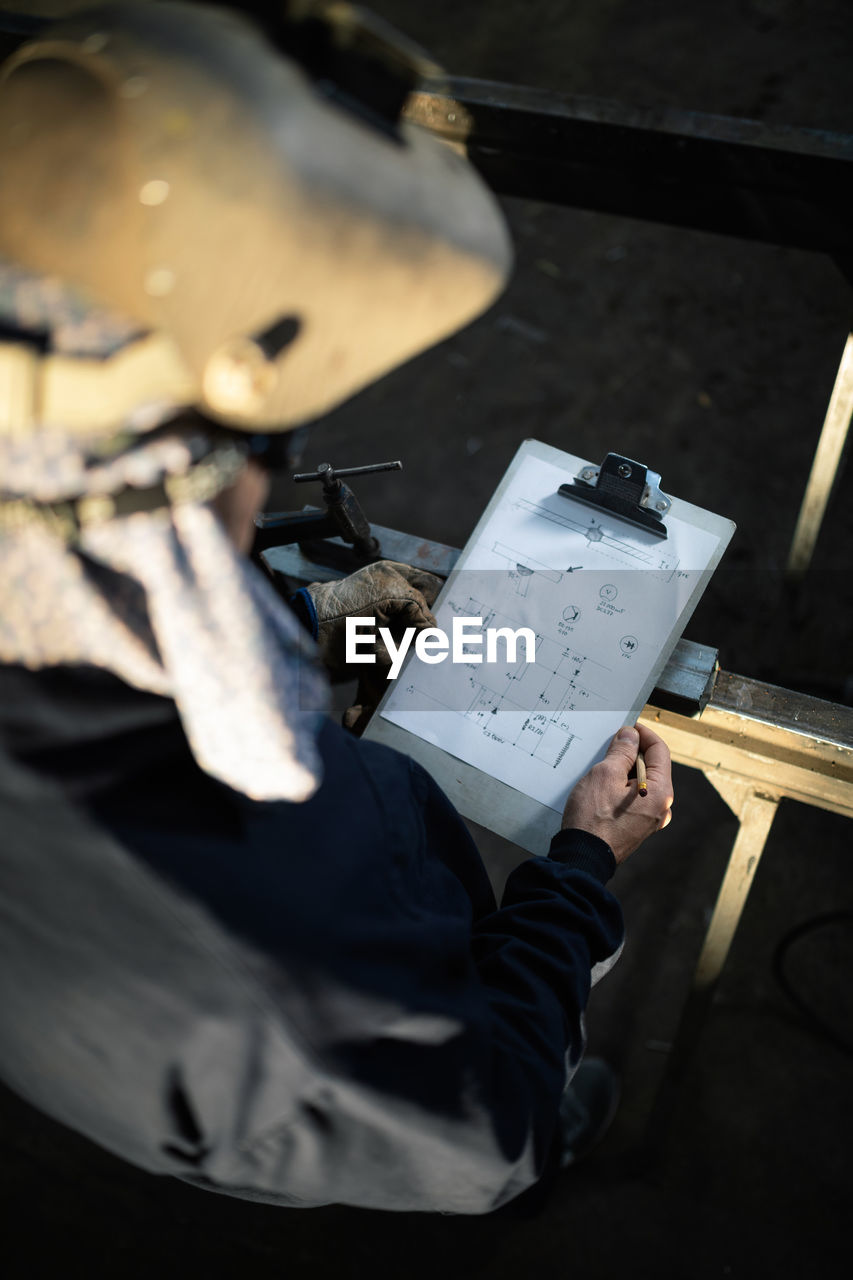 Male worker in hardhat and overall taking notes in notebook with pen on table near metal constructions in light garage