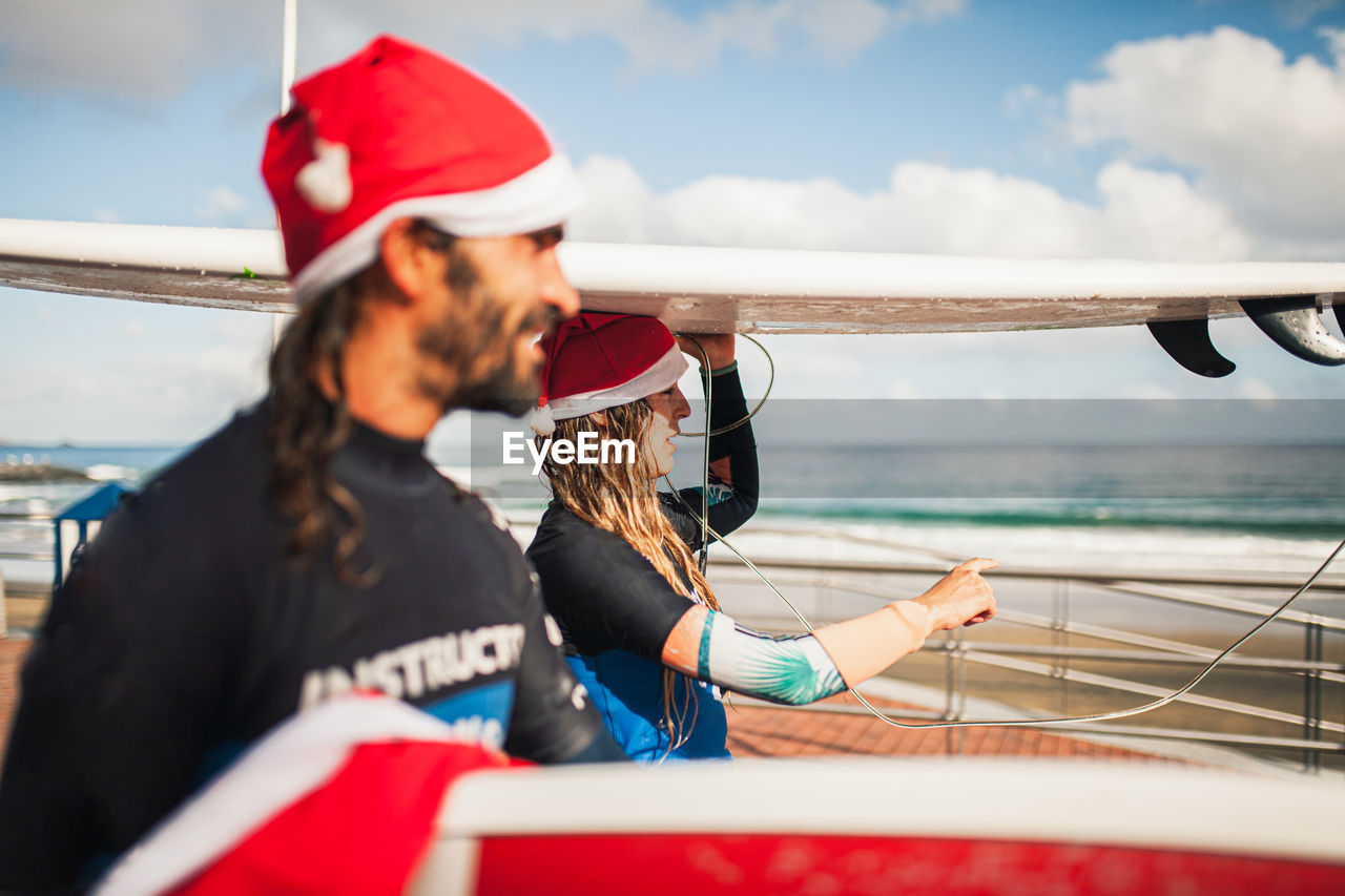 Side view of couple wearing santa hats carrying surfboards on heads