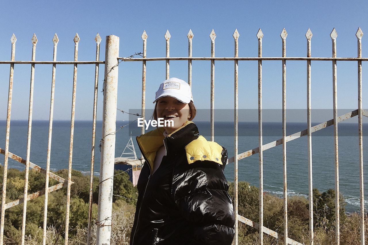 fence, one person, protection, security, nature, sky, standing, clothing, day, leisure activity, waist up, looking at camera, portrait, men, lifestyles, adult, three quarter length, outdoors, architecture, person, hat, smiling, metal, front view, casual clothing, sea, emotion, water, sports, human face, land, railing