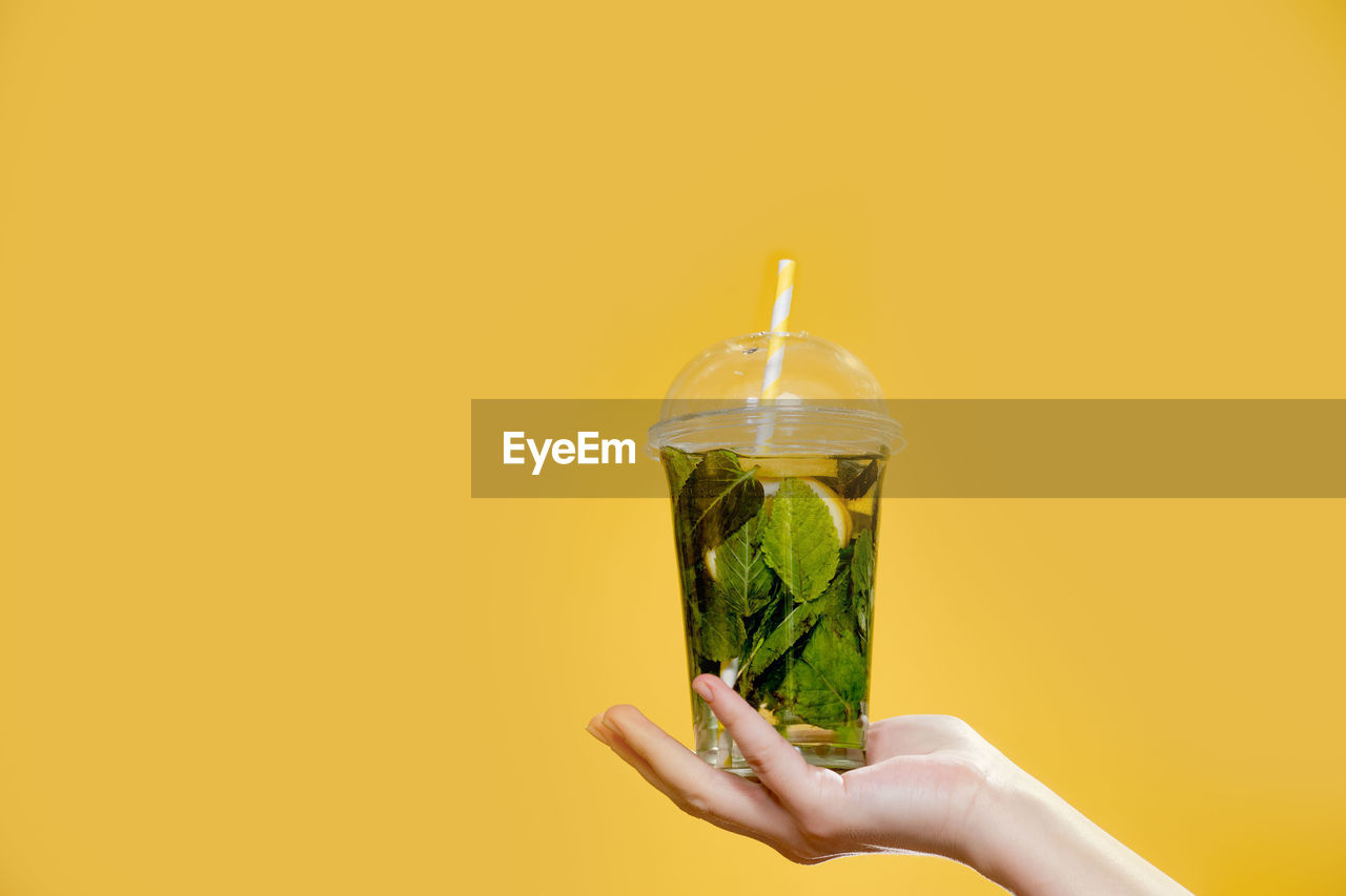 CLOSE-UP OF HAND HOLDING GLASS OF YELLOW OVER BACKGROUND