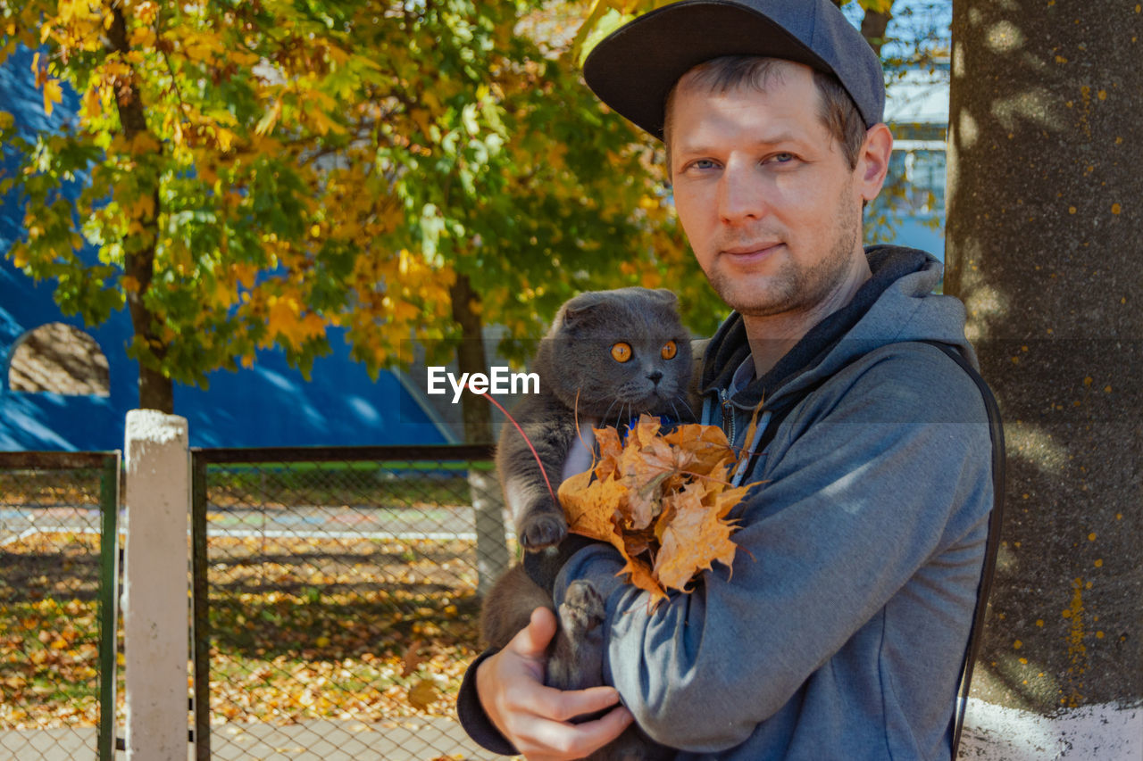 autumn, one person, animal, animal themes, portrait, looking at camera, men, adult, nature, mammal, one animal, domestic animals, holding, pet, day, hat, emotion, clothing, person, plant, leaf, tree, outdoors, smiling, waist up, casual clothing, leisure activity, plant part, flower, front view, lifestyles, child, human face, animal wildlife, fence, standing, cap, happiness