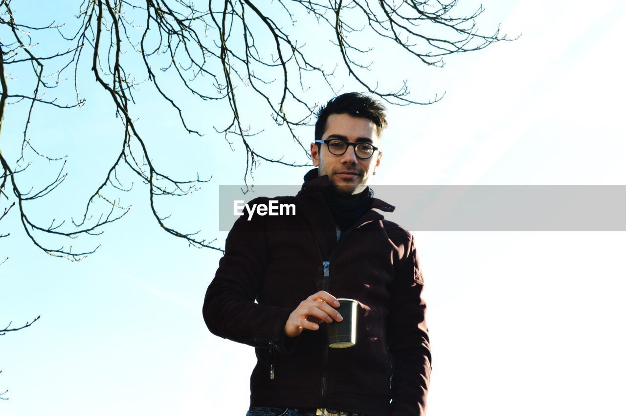 Handsome young man holding coffee mug while standing against clear sky during winter