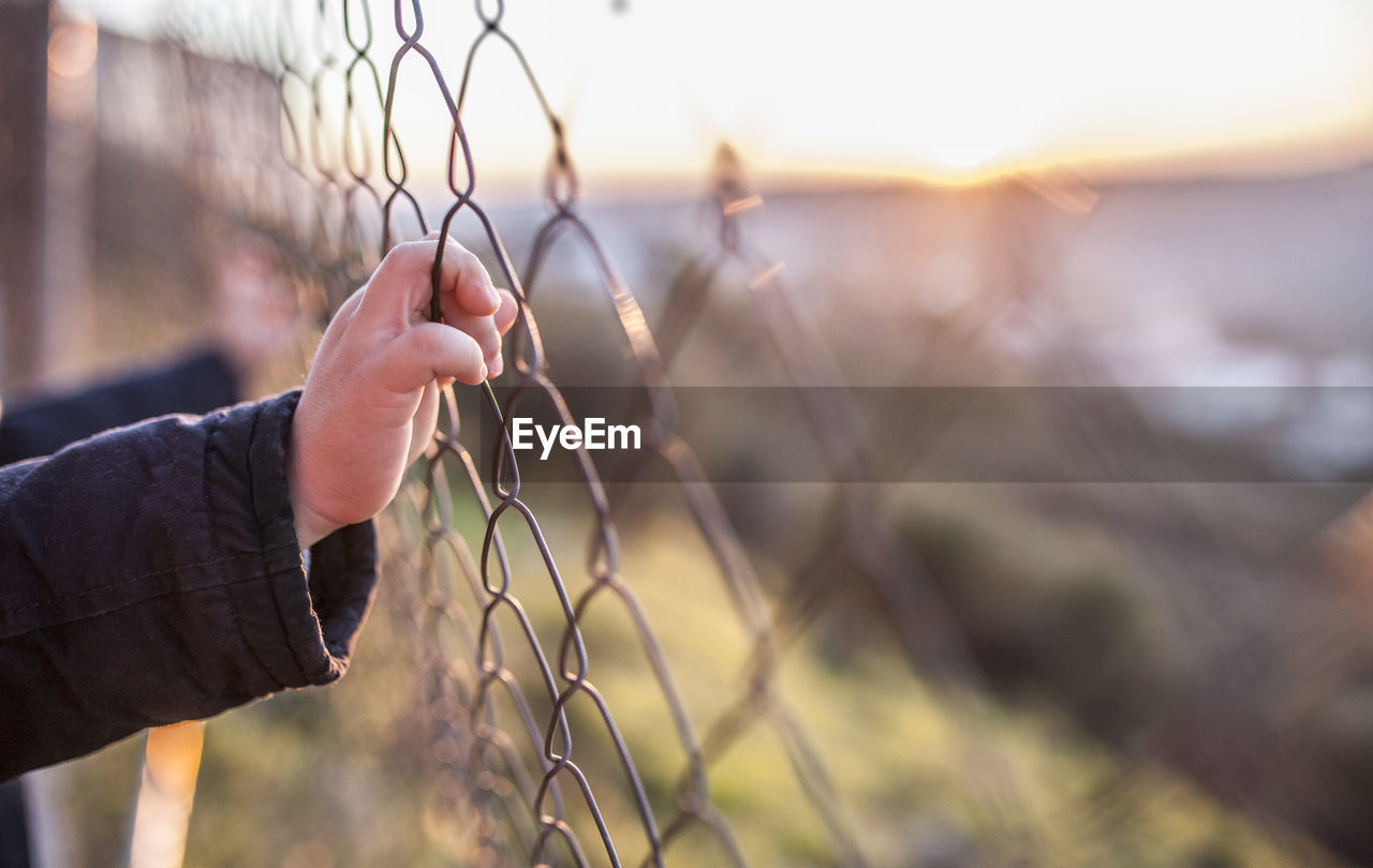 Kid boy holding hands on wire fence at sunset