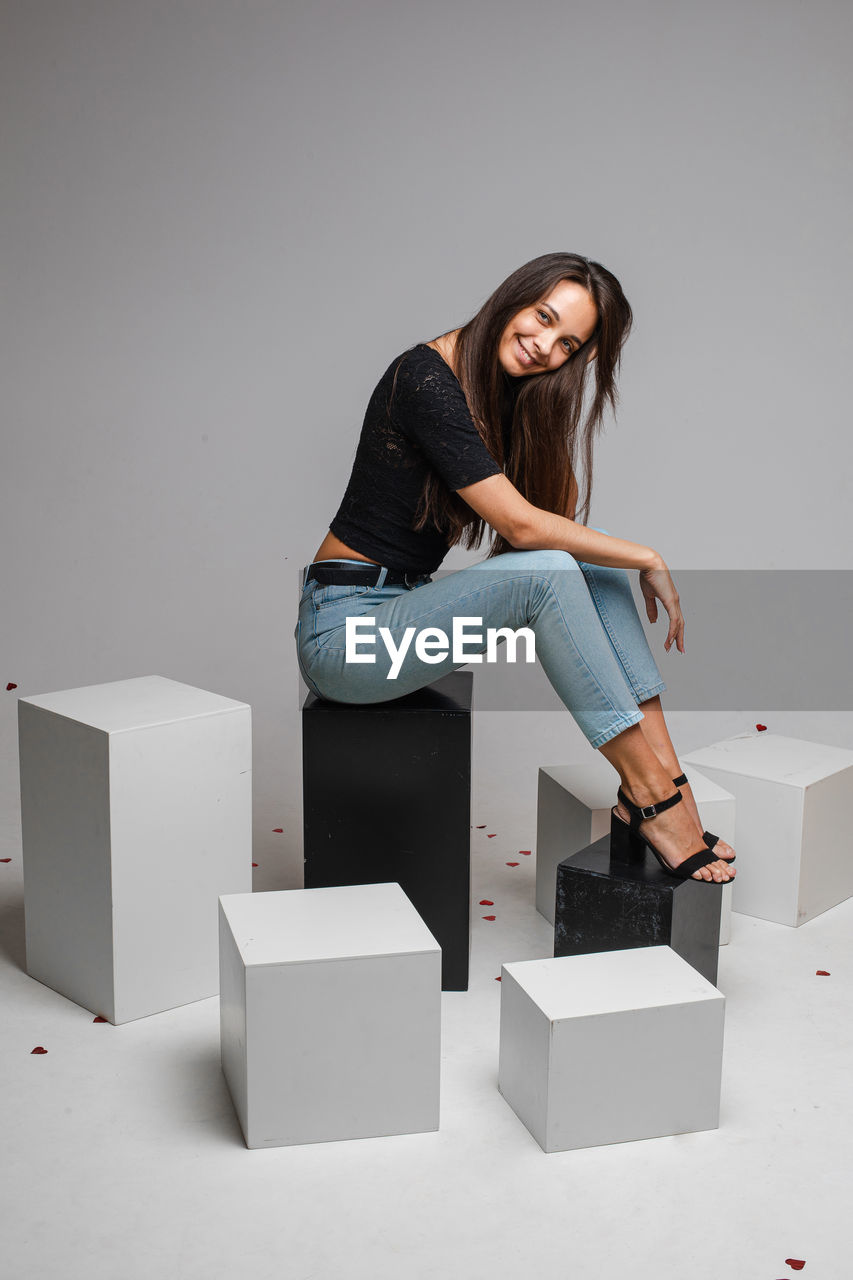 Smiling pretty young woman posing in studio while sitting on black boxes, isolated on grey 