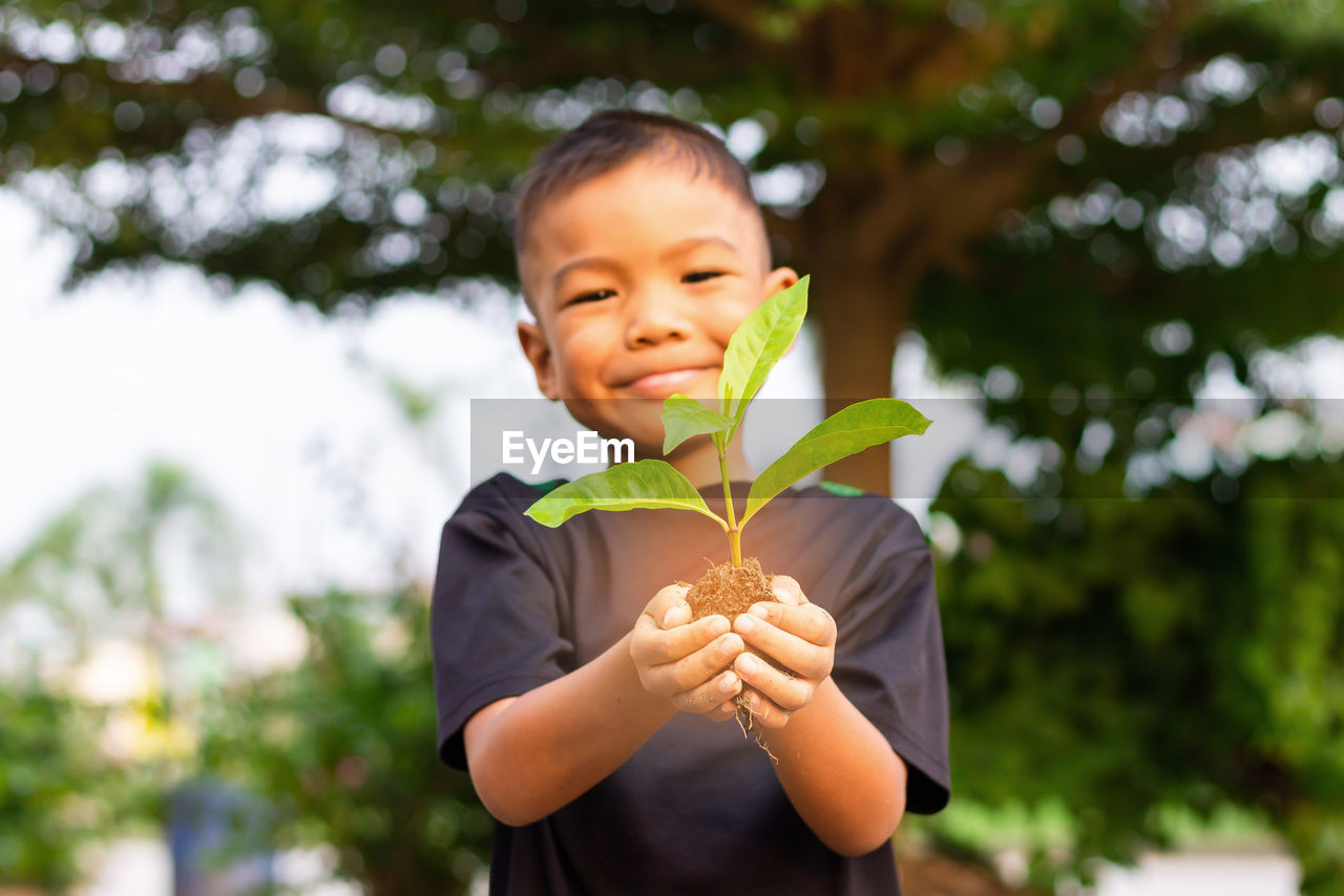 Low angle portrait of boy holding sapling while standing against trees