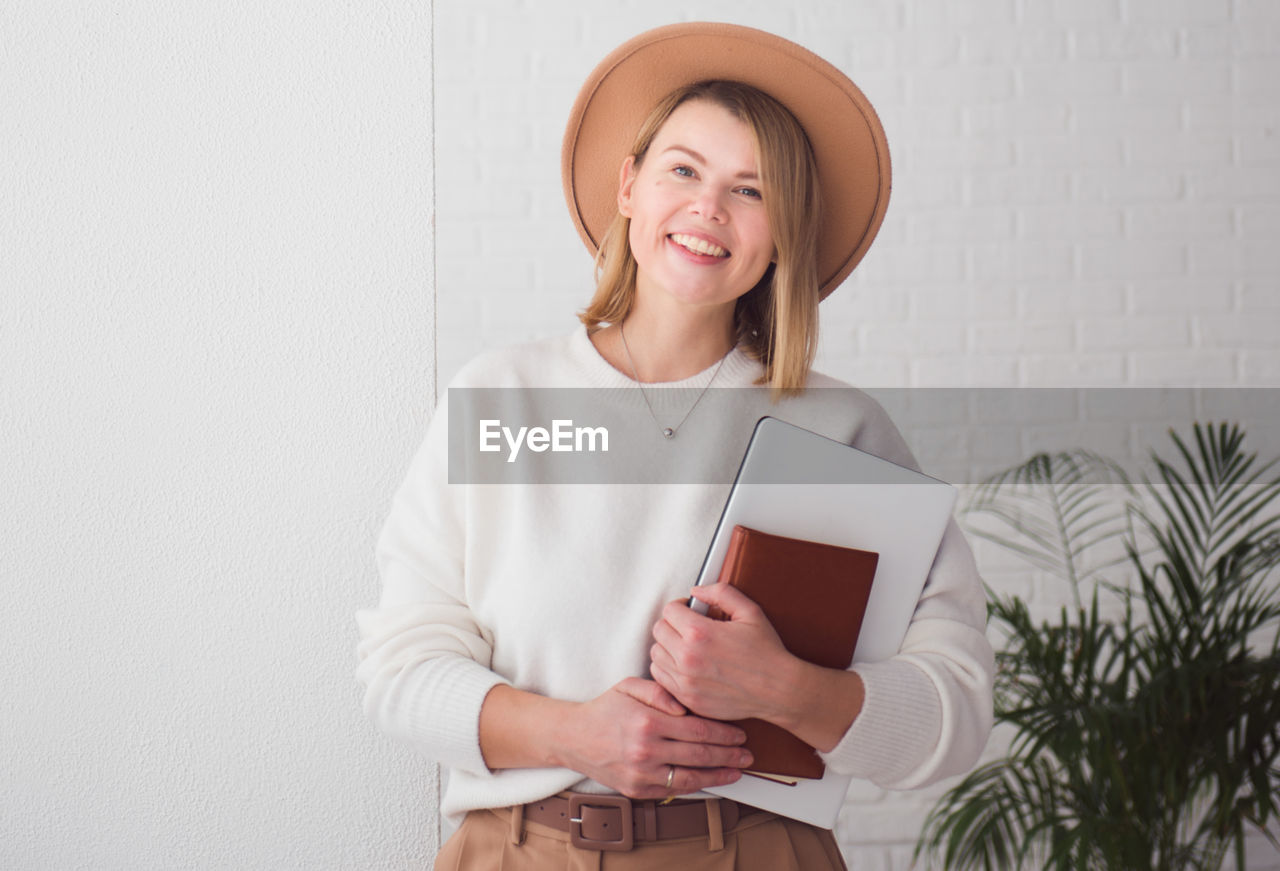 Trendy woman freelancer wearing brown hat holding notebook, smiling and looking at camera