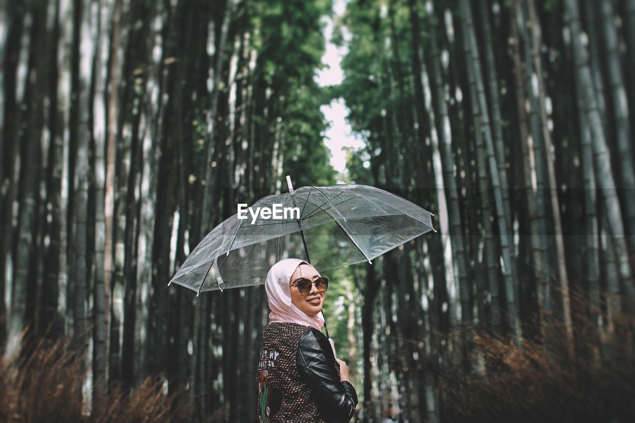 Smiling woman holding umbrella while standing against trees