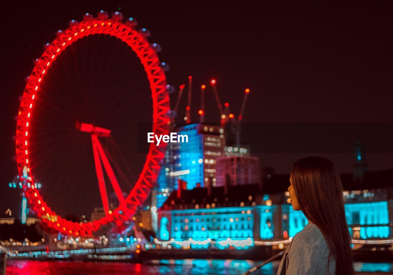 Woman looking at illuminated ferris wheel against sky at night