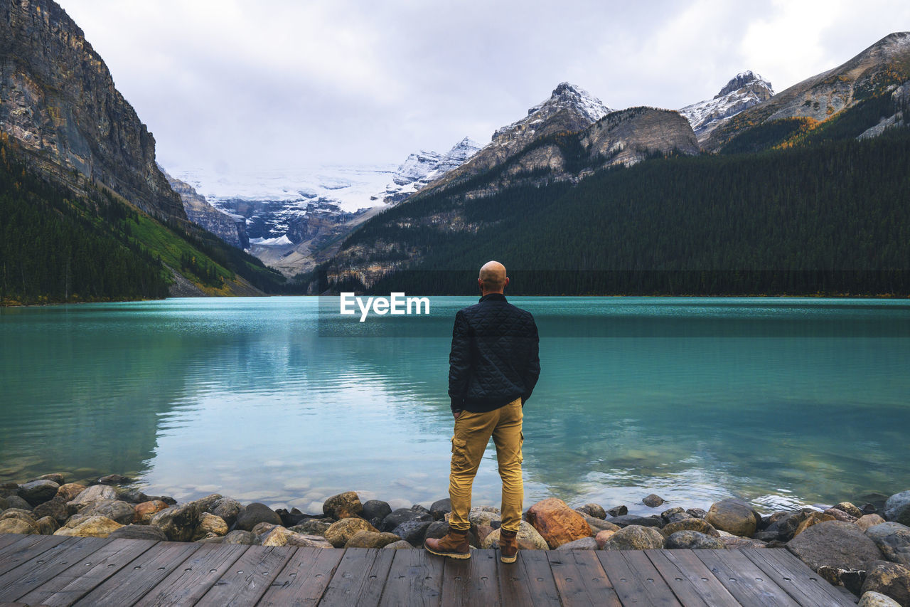 Rear view of man standing on jetty looking at lake and mountains