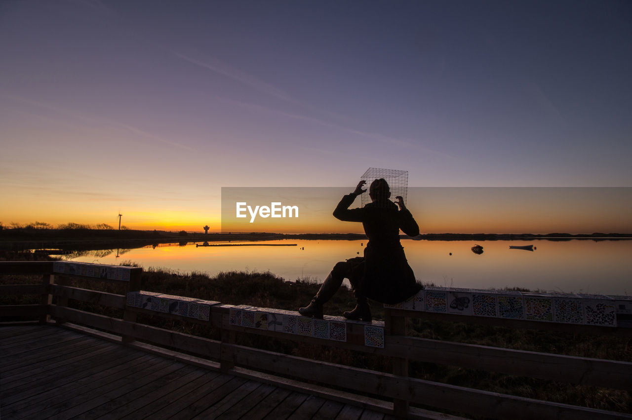 Silhouette of man sitting on railing by sea