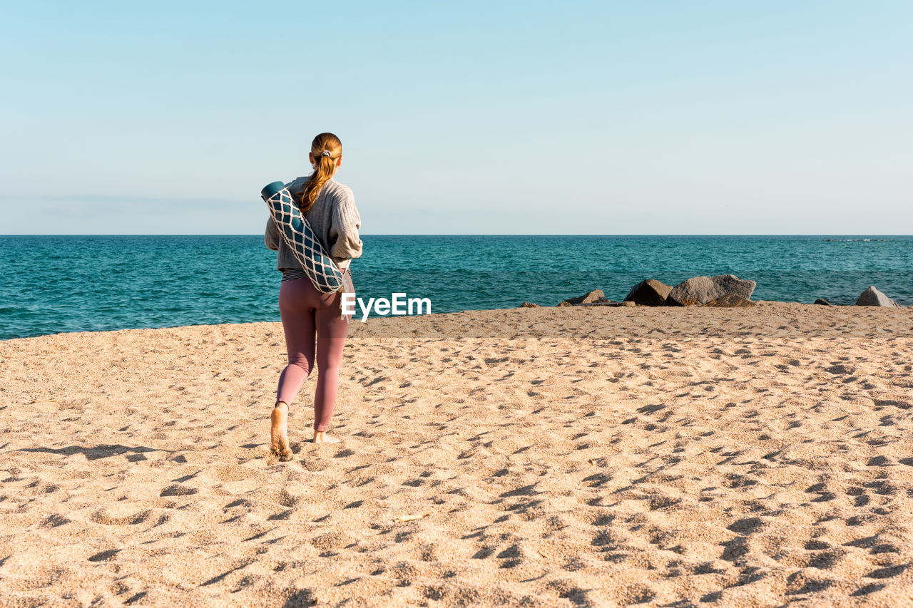 Full body back view of unrecognizable of young barefoot female in activewear with rolled yoga mat looking at distance while standing on sandy beach near sea